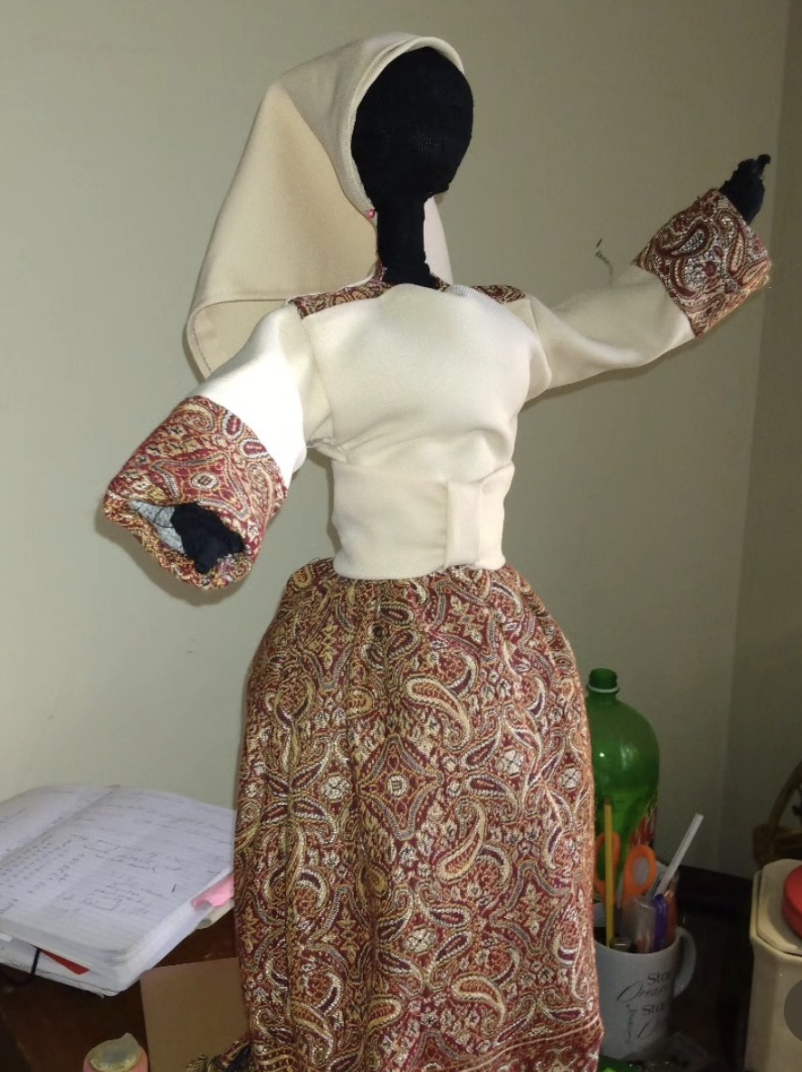 A handmade soft-sculpture doll with a black head, cream torso, and burgundy patterned fabric arms and skirt, wearing a cream headwrap